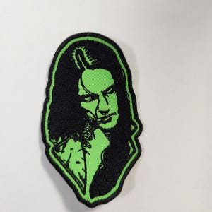 May include: An embroidered patch featuring a stylized portrait of a person with long dark hair. The design uses a high-contrast color scheme of vibrant lime green and deep black, with a thick black border outlining the irregular shape of the patch.