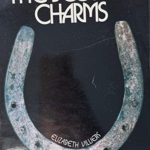 May include: A book titled The Book of Charms by Elizabeth Villiers with a black cover featuring a large, weathered, metallic horseshoe in the centre. The title is printed in white, stylised lettering at the top, and the author's name is at the bottom.