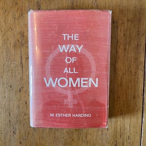 Puede incluir: Un libro rojo con el título "The Way of All Women" en texto blanco. El libro tiene un símbolo femenino en la portada y el nombre del autor, M. Esther Harding, está impreso en la parte inferior.
