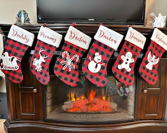 May include: Six Christmas stockings with red and black buffalo plaid patterns hang above a fireplace. Each stocking has a white cuff with a name embroidered in red. The stockings feature holiday-themed designs.