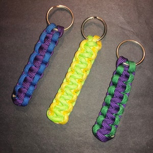May include: Three handmade paracord keychains in blue and purple, yellow and green, and purple and green. Each keychain features a woven design and a silver-colored metal keyring. The keychains are displayed on a dark surface.