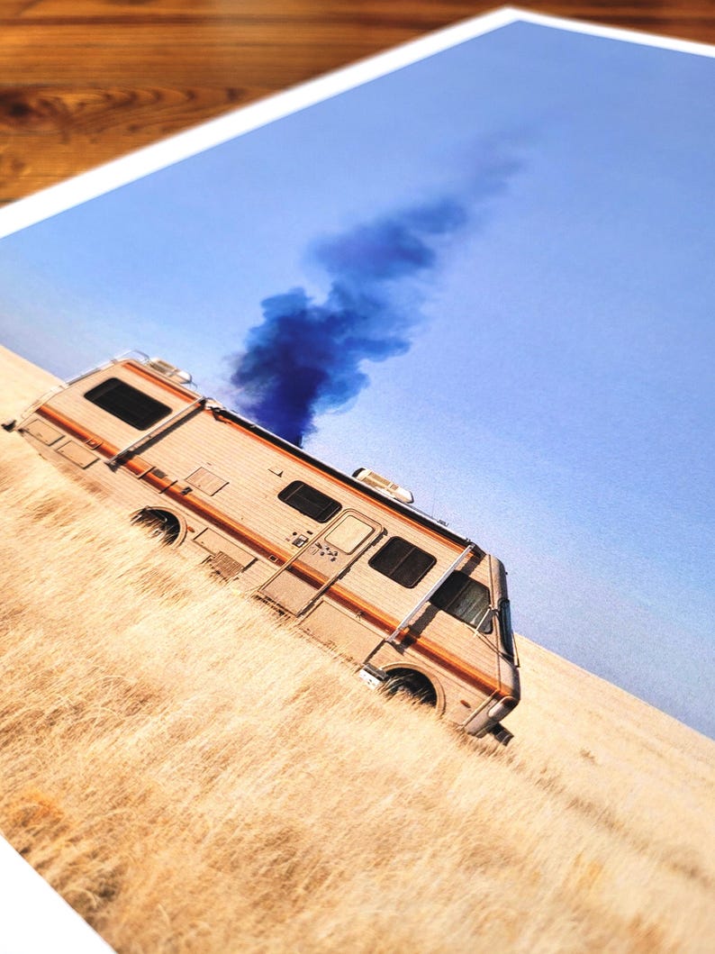 May include: A white and brown recreational vehicle with a blue smoke plume coming from the back, parked in a field of dry grass under a blue sky.