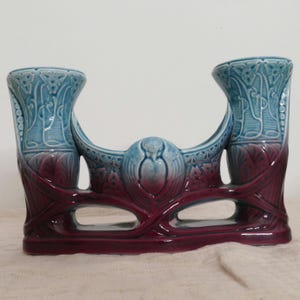 Może przedstawiać: Ceramiczny świecznik z dwoma wazonami o gradientowym kolorze, przechodzącym od głębokiego burgundu u podstawy do jasnego błękitu u góry. Przedmiot posiada skomplikowane, formowane wzory reliefowe oraz dwa oddzielne cylindryczne otwory.