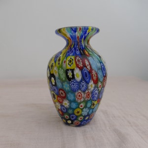 Puede incluir: Un pequeño jarrón de vidrio con un vibrante patrón millefiori que presenta numerosos motivos florales coloridos en tonos rojo, azul, amarillo, verde y blanco. El jarrón tiene un cuerpo redondeado y un borde acampanado con densas varillas de vidrio.