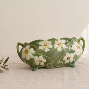 May include: An oval ceramic planter with a glossy sage green glaze, featuring raised white daffodil motifs with yellow centers. The vessel has two arched handles on the sides, a scalloped rim, and stands on four small, integrated feet.