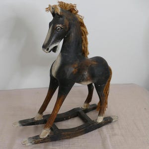 May include: A wooden rocking horse featuring a dark brown and black painted finish with a white blaze on the face. The horse has a tan yarn mane and tail, and is mounted on two curved wooden rockers connected by two horizontal crossbars.