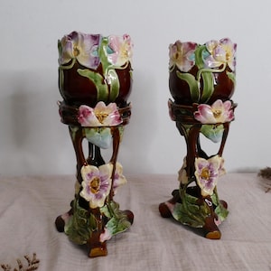 May include: A pair of tall ceramic vases featuring a deep brown glaze with three-dimensional sculpted flowers in shades of pink, purple, and white, accented by green leaves. The vases have a cylindrical top section supported by a branched base.