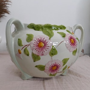Puede incluir: Un cuenco de cerámica de color crema con dos asas y tres patas pequeñas. El exterior presenta flores rosas pintadas en relieve tridimensional con centros amarillos y hojas verdes que envuelven el cuerpo del recipiente.
