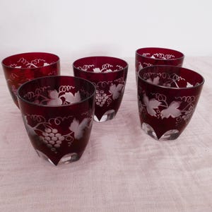 Könnte beinhalten: Ein Set aus vier tiefroten Glastumblern mit einem klaren, geätzten Design aus Weintrauben und Weinblättern. Jeder Becher verfügt über einen dicken, klaren Glasboden und einen glatten, abgerundeten Rand mit durchgehendem, mattiertem Muster.
