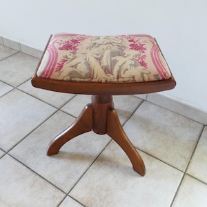 Puede incluir: Un taburete de madera con asiento curvo tapizado en tela beige con un patrón de tapiz floral y de querubines en tonos rosa y crema. El taburete cuenta con una base central que se divide en tres patas de madera curvadas.