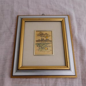 May include: A framed artwork featuring a small rectangular illustration of a city skyline with a yellow sun and clouds above a body of water. The frame has a gold and silver finish with a light beige matting surrounding the central image.