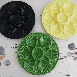 May include: Three circular ceramic oyster plates in black, yellow, and green. Each plate features six shell-shaped indentations arranged around a central circular compartment, with a textured basket-weave pattern on the outer rim.