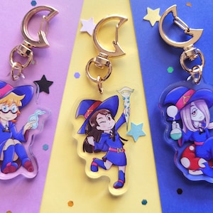 May include: Three colorful acrylic keychains featuring anime characters with witch hats. The characters are holding wands, a potion bottle, and a book. Each keychain has a gold metal crescent moon-shaped clasp.