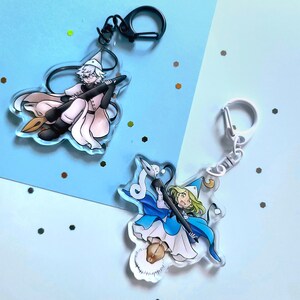 May include: Two acrylic keychains featuring anime-style characters. One keychain has a character in a pink outfit with a black broom. The other keychain has a character in a blue outfit with a black broom. Both keychains have a silver or black clasp.