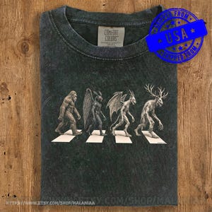 Op de afbeelding: Een antracietgrijs acid-wash T-shirt met een afbeelding van vier cryptiden die in een rij over witte strepen lopen. De wezens zijn Bigfoot, Mothman, een gevleugelde demon en een jackalope. Het label aan de binnenkant vermeldt Comfort Colors.