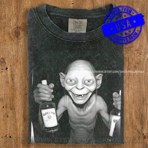 May include: Dark gray t-shirt featuring a graphic of Gollum from Lord of the Rings holding two bottles of liquor. The shirt has a distressed look and a Comfort Colors tag. A blue stamp indicates it was shipped from the USA.
