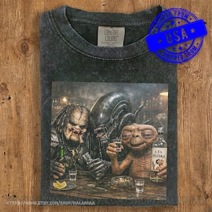 Pode incluir: Uma t-shirt cinzento-carvão com lavagem ácida, apresentando uma estampa gráfica de um Predator, um Alien e o E.T. sentados num bar com bebidas. A t-shirt tem uma etiqueta Comfort Colors no pescoço. O gráfico mostra as personagens com copos e uma garrafa com o rótulo E.T. Palinka.