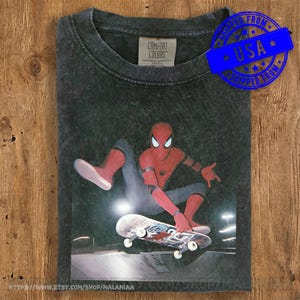 May include: A charcoal grey acid-wash t-shirt featuring a graphic of a red and blue superhero character performing a skateboard trick at night. The shirt has a Comfort Colors brand label at the inner neckline and a crew neck collar design.
