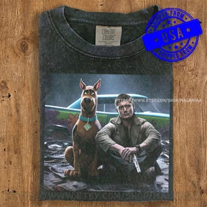 May include: Dark gray Comfort Colors t-shirt featuring a graphic of Scooby-Doo and a man with a gun. Scooby-Doo is brown with a blue collar. The man is wearing a green jacket. The shirt has a blue "USA" stamp.