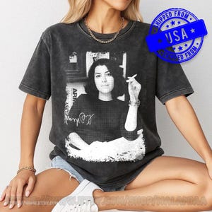 May include: A charcoal grey short-sleeve t-shirt featuring a black and white graphic print of a person holding a cigarette. The design includes a signature and a distressed, vintage-style wash. The shirt has a crew neckline and a relaxed fit.