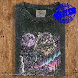 Può includere: Una t-shirt girocollo grigio antracite con una stampa grafica raffigurante un gatto soffice con occhiali da sole rotondi e una corona, una luna piena, un dinosauro blu, una cascata e un'aurora boreale su un paesaggio montano. L'etichetta riporta la scritta Comfort Colors.