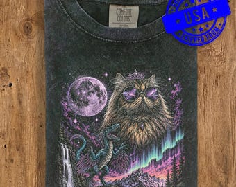 May include: A charcoal gray crew neck t-shirt with a graphic print featuring a fluffy cat wearing round sunglasses and a crown, a full moon, a blue dinosaur, a waterfall, and an aurora borealis over a mountain landscape. The label reads Comfort Colors.
