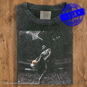 May include: Dark gray Comfort Colors t-shirt featuring a graphic of Darth Vader dunking a basketball. The image is set against a basketball court backdrop. A blue "Shipped From USA" sticker is on the top right. The shirt has a vintage, distressed look.