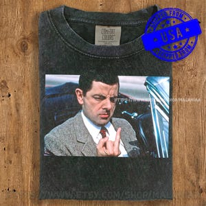 May include: Dark gray Comfort Colors t-shirt featuring a graphic of a man in a suit giving the middle finger. The image is a close-up of the man's face. The shirt has a vintage, worn look.