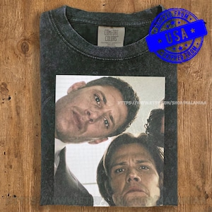 May include: Dark gray Comfort Colors t-shirt featuring a graphic of two men looking down. The graphic is a close-up of their faces. The t-shirt has a crew neck and a vintage wash. The text "USA" is on a blue sticker.