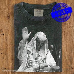 Può includere: T-shirt Comfort Colors grigio scuro con un'immagine in bianco e nero di una figura religiosa che alza una mano. La figura indossa una veste bianca e tiene in mano un libro. La maglietta ha un aspetto vintage.