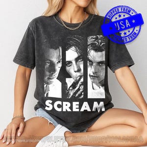 Puede incluir: Una camiseta de manga corta en color gris carbón con un estampado gráfico en blanco y negro de tres rostros de la película Scream. La palabra SCREAM está impresa en letras mayúsculas blancas grandes en la parte inferior. La tela tiene un aspecto desgastado.