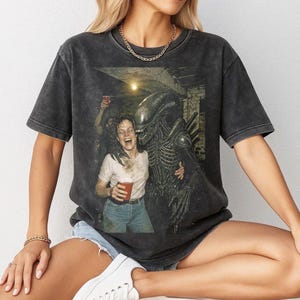 Puede incluir: Camiseta gris oscuro con un gráfico de la película "Alien". La imagen muestra a una mujer riendo, abrazada por el Xenomorfo. La camiseta tiene un aspecto vintage y desgastado.