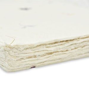 May include: A stack of off-white handmade paper with a rough, deckled edge. The paper has a fibrous, textured surface with small embedded dark specks and a single visible blue petal fragment protruding from the side of the layered stack.