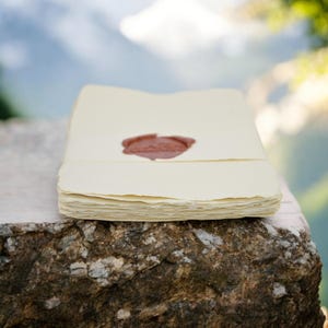 May include: A stack of blank, cream-colored paper with a red wax seal on the top sheet. The paper is sitting on a rough, brown rock.