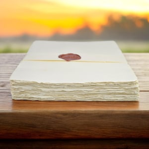 May include: A stack of blank, white paper sheets with a red wax seal on the top sheet. The paper is on a wooden table with a blurred background of a sunset.