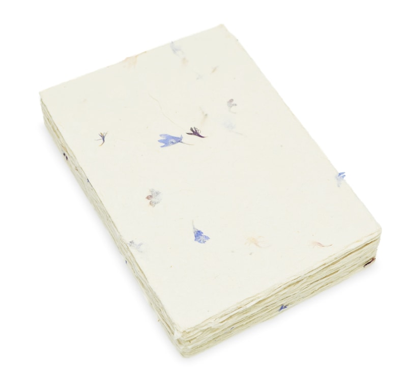 May include: A rectangular stack of off-white handmade paper sheets with a deckled edge. The paper features embedded small, dried blue and purple flower petals scattered across the surface, creating a textured and organic appearance throughout.