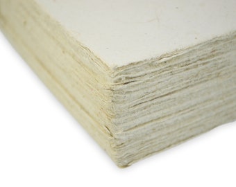 May include: A stack of thick, off-white handmade paper sheets with visible fibrous texture and rough, deckled edges. The paper has a cream-colored hue and a layered appearance, showing the individual thickness of each sheet in the stack.