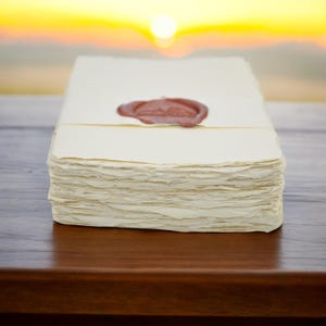 May include: A stack of cream-colored handmade paper with deckled edges, sealed with a brown wax stamp. The paper is stacked on a dark wooden surface, with a blurred sunset in the background. The paper appears to be for letters or invitations.