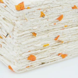May include: A stack of handmade paper with a rough, textured surface. The paper is off-white with small orange and yellow flecks throughout. The edges of the paper are uneven, giving it a rustic appearance. The paper is likely used for crafts or stationery.