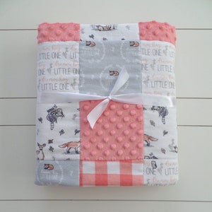May include: A pink and gray baby blanket with a patchwork design. The blanket features a variety of fabrics, including a pink minky fabric, a gray fabric with a white floral pattern, and a white fabric with a pink and white gingham pattern. The blanket is tied with a white ribbon and has the text "Little One" repeated on it.