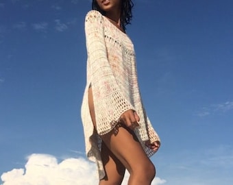 Puede incluir: Una mujer con un top de crochet blanco de corte holgado y mangas largas, de pie en una playa de arena con un cielo azul y nubes blancas de fondo.