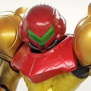 Könnte beinhalten: Detaillierte Actionfigur einer Figur in einem gold-roten Anzug. Die Figur hat einen roten Helm mit grünem Visier, goldene Schulterpolster und eine blaue Waffe. Die Figur ist in einer dynamischen Haltung dargestellt.