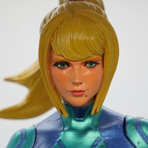 Puede incluir: Figura detallada de un personaje femenino con cabello rubio recogido en una cola de caballo. Tiene ojos verdes y viste un traje azul y verde. La figura es un objeto de colección, probablemente de un videojuego o serie de cómics.