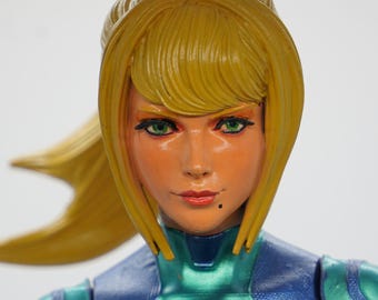 May include: A detailed figurine of a female character with blonde hair styled in a ponytail. She has green eyes, orange skin, and is wearing a teal and blue suit. The figurine is a collectible item, likely from a video game or comic book series.
