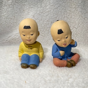 Puede incluir: Dos pequeñas figuritas de cerámica de niños con ropa tradicional china. El niño de la izquierda lleva una camisa amarilla y pantalones azules, mientras que el niño de la derecha lleva una camisa azul y pantalones rosas. Ambas figuritas tienen el pelo negro y están sentados con las piernas cruzadas.