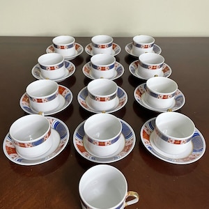 Puede incluir: Un juego de 13 tazas de té y platillos blancos con un patrón geométrico azul, rojo y dorado. Las tazas y los platillos están dispuestos en círculo sobre una mesa de madera.