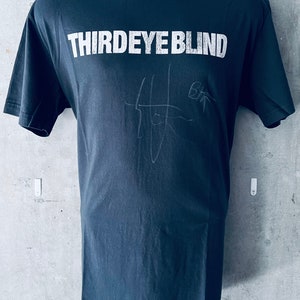May include: A dark gray t-shirt with the band name "THIRDEYE BLIND" printed in white on the front. The shirt is signed in white ink.