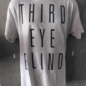 May include: White t-shirt with the text "THIRD EYE BLIND" printed in bold, black capital letters. The shirt is displayed on a black mannequin and has short sleeves.
