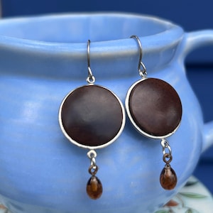 May include: A pair of drop earrings featuring circular dark brown wooden discs set in silver-toned metal frames. Each earring hangs from a silver-toned hook and includes a small, translucent amber-colored teardrop-shaped bead dangling below.