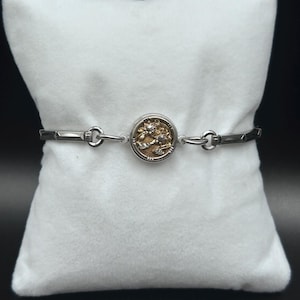 May include: A silver-toned bracelet featuring a central circular charm with a textured gold-colored relief design. The chain consists of alternating smooth, elongated rectangular links and small circular jump rings, creating a balanced, metallic aesthetic.